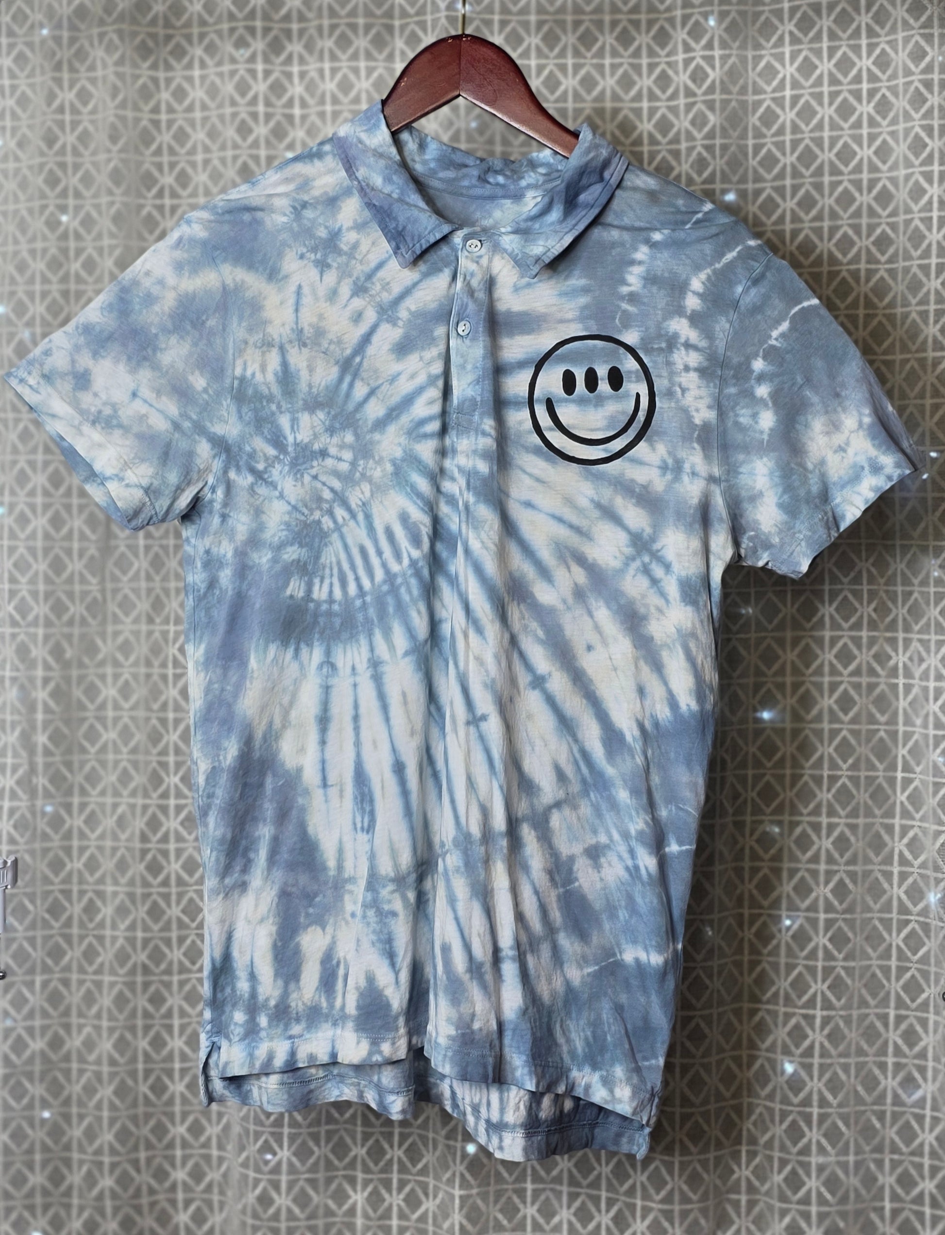 Tie-dye polo shirt with smiley face design on a hanger against a patterned wall.