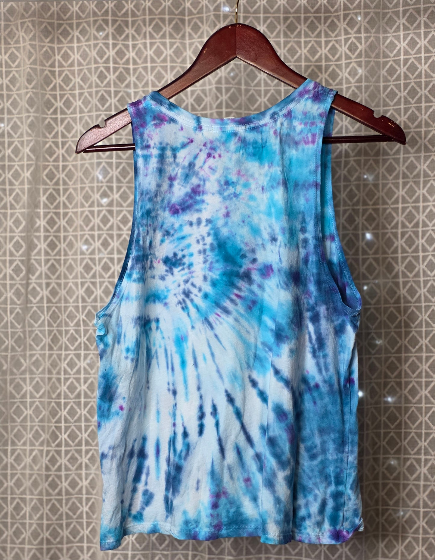 Tie-dye tank top on a hanger against a patterned wall.