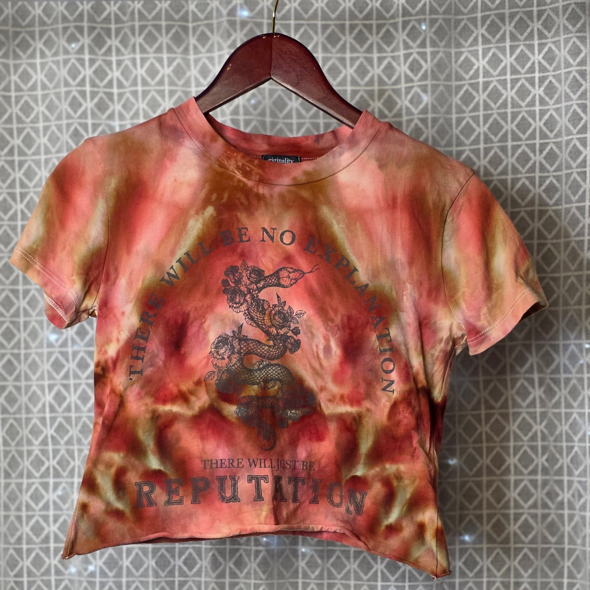 Tie-dye t-shirt with text and graphic design on a hanger against a patterned wall.