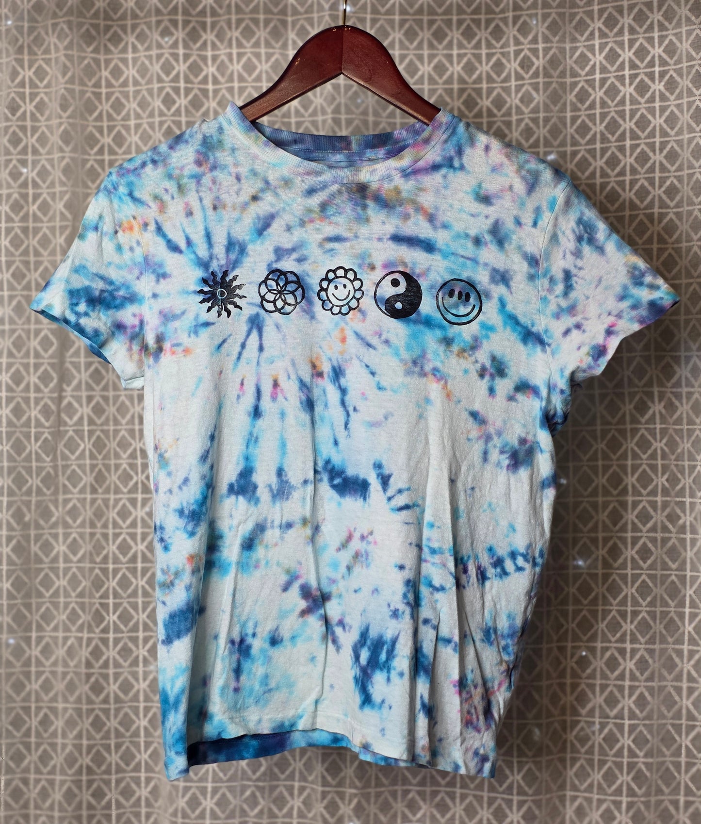 Tie-dye t-shirt with symbols on a hanger against a patterned wall.