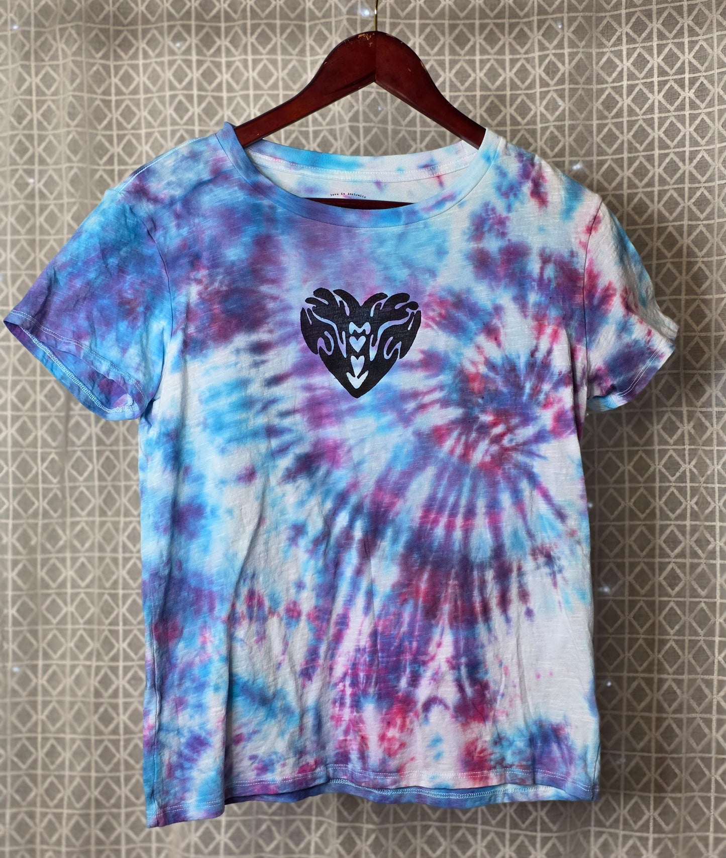 Tie-dye t-shirt with a black heart design on a hanger against a patterned wall.
