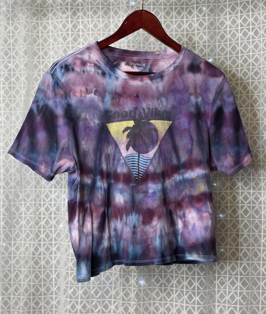Tie-dye t-shirt with a logo on a hanger against a patterned wall.