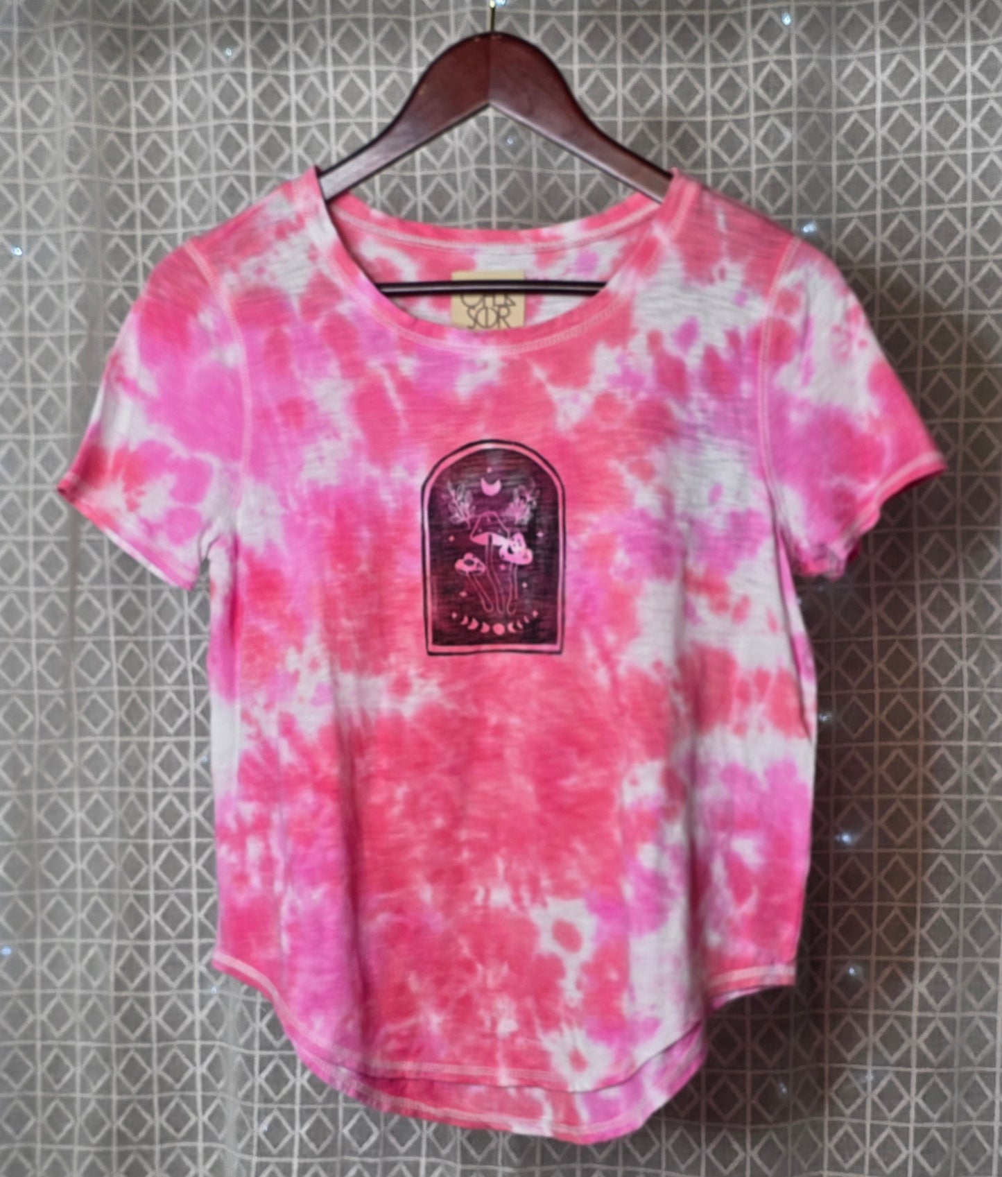 Pink and white tie-dye t-shirt with a graphic patch on a hanger against a patterned wall.