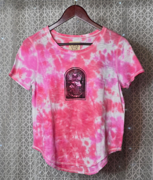 Pink and white tie-dye t-shirt with a graphic patch on a hanger against a patterned wall.
