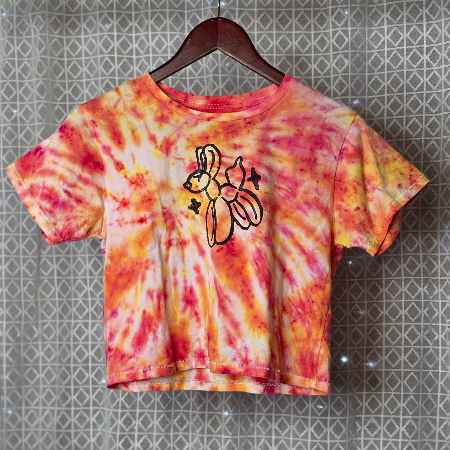 Tie-dye t-shirt with a balloon animal print on a hanger against a patterned wall.