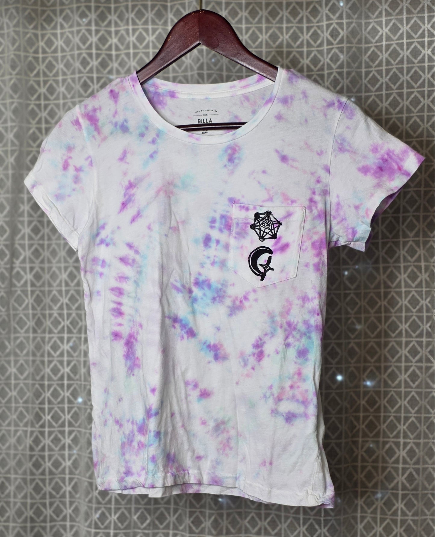 White t-shirt with purple and pink moon pattern on a hanger against a textured wall.