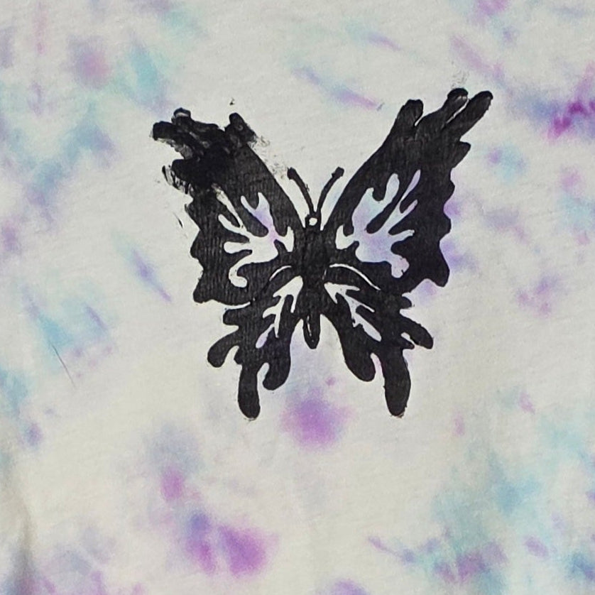Tie-dye t-shirt with a butterfly design on a hanger against a patterned wall.