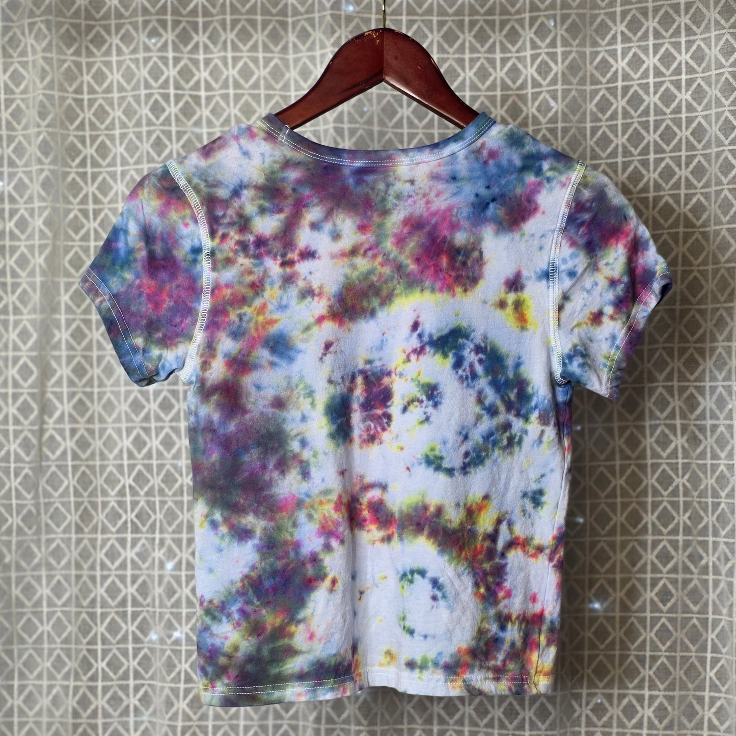 Colorful tie-dye t-shirt on a hanger against a patterned wall.