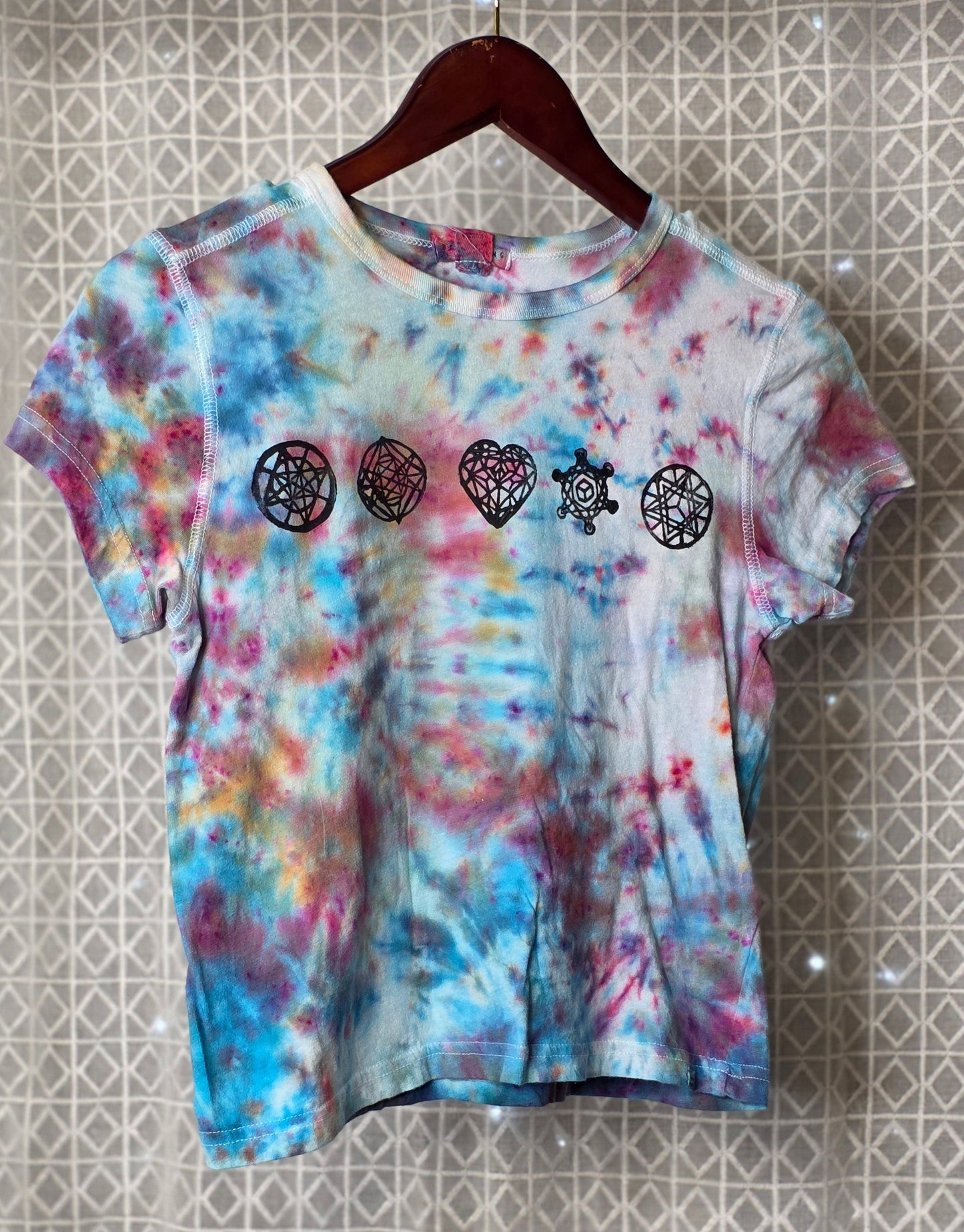 Tie-dye t-shirt with heart and star designs on a hanger against a patterned wall.