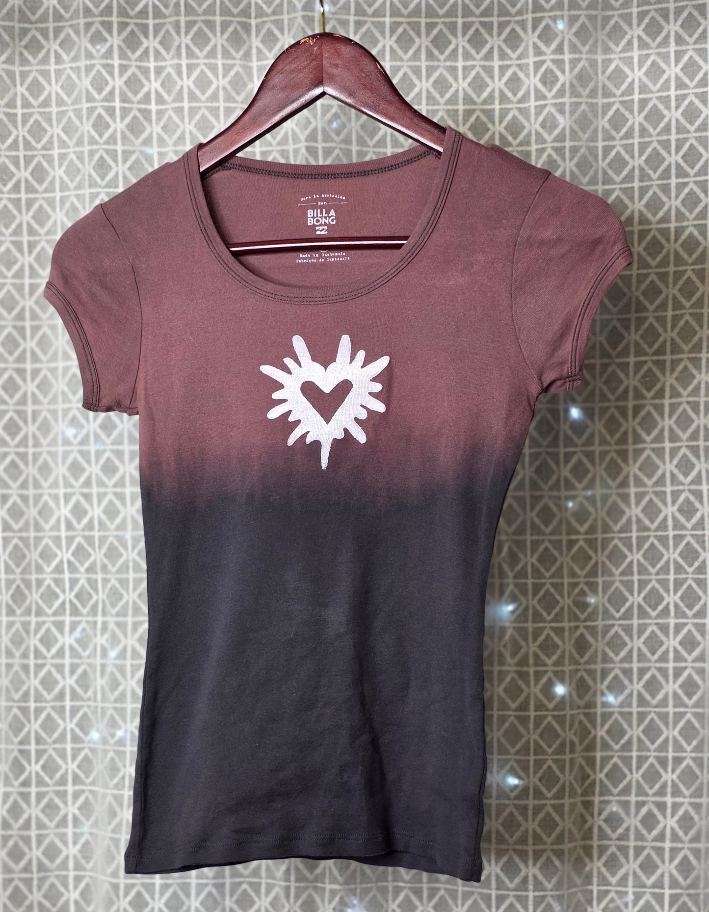 Gradient t-shirt with a white heart and sun design on a hanger against a patterned wall.