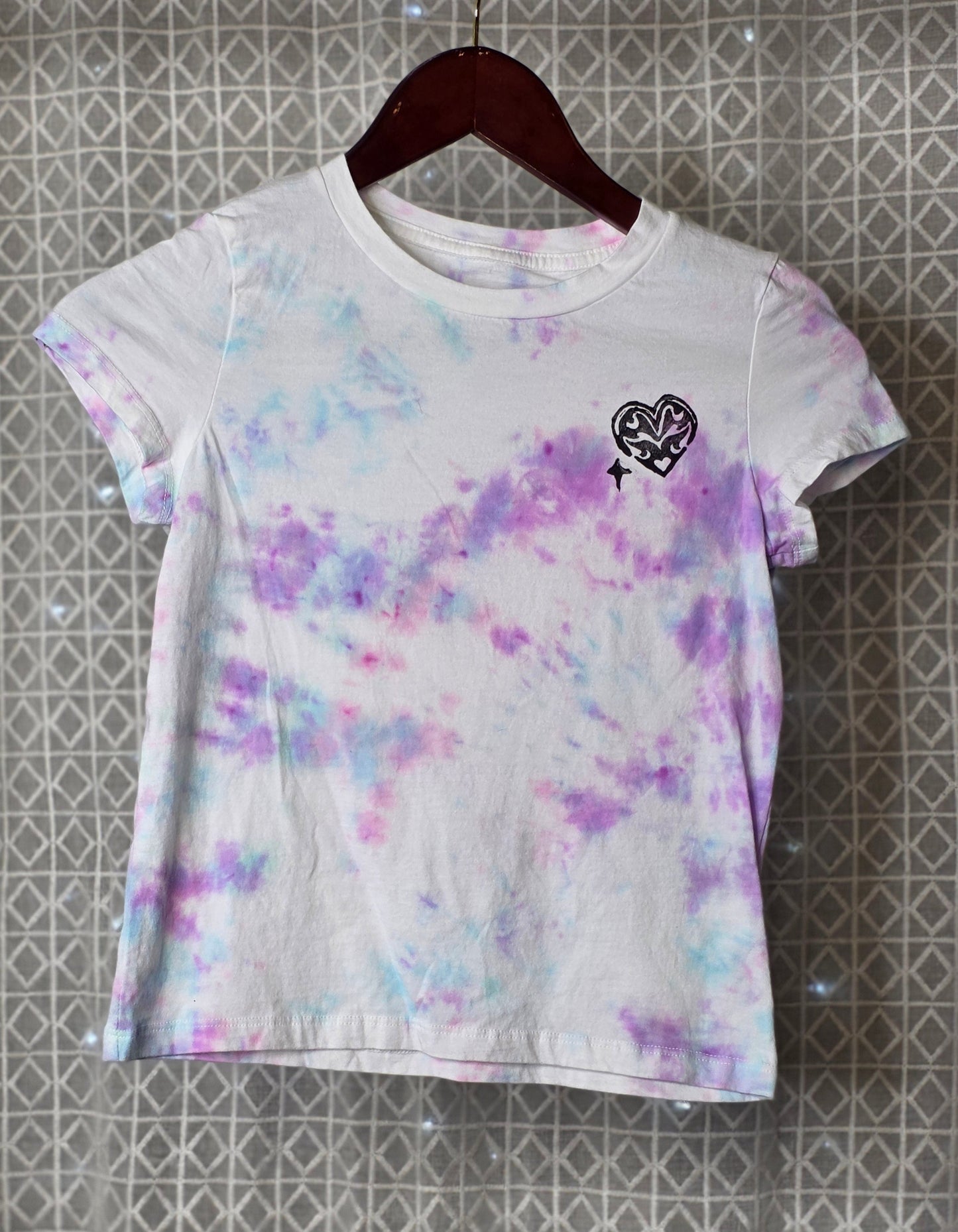 Tie-dye t-shirt with a heart design on a hanger against a patterned wall.
