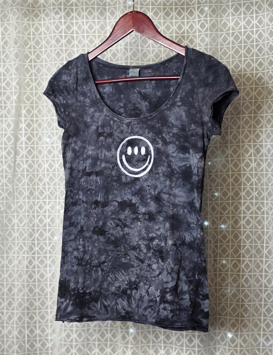 Black tie-dye t-shirt with a white smiley face design on a hanger against a patterned wall.