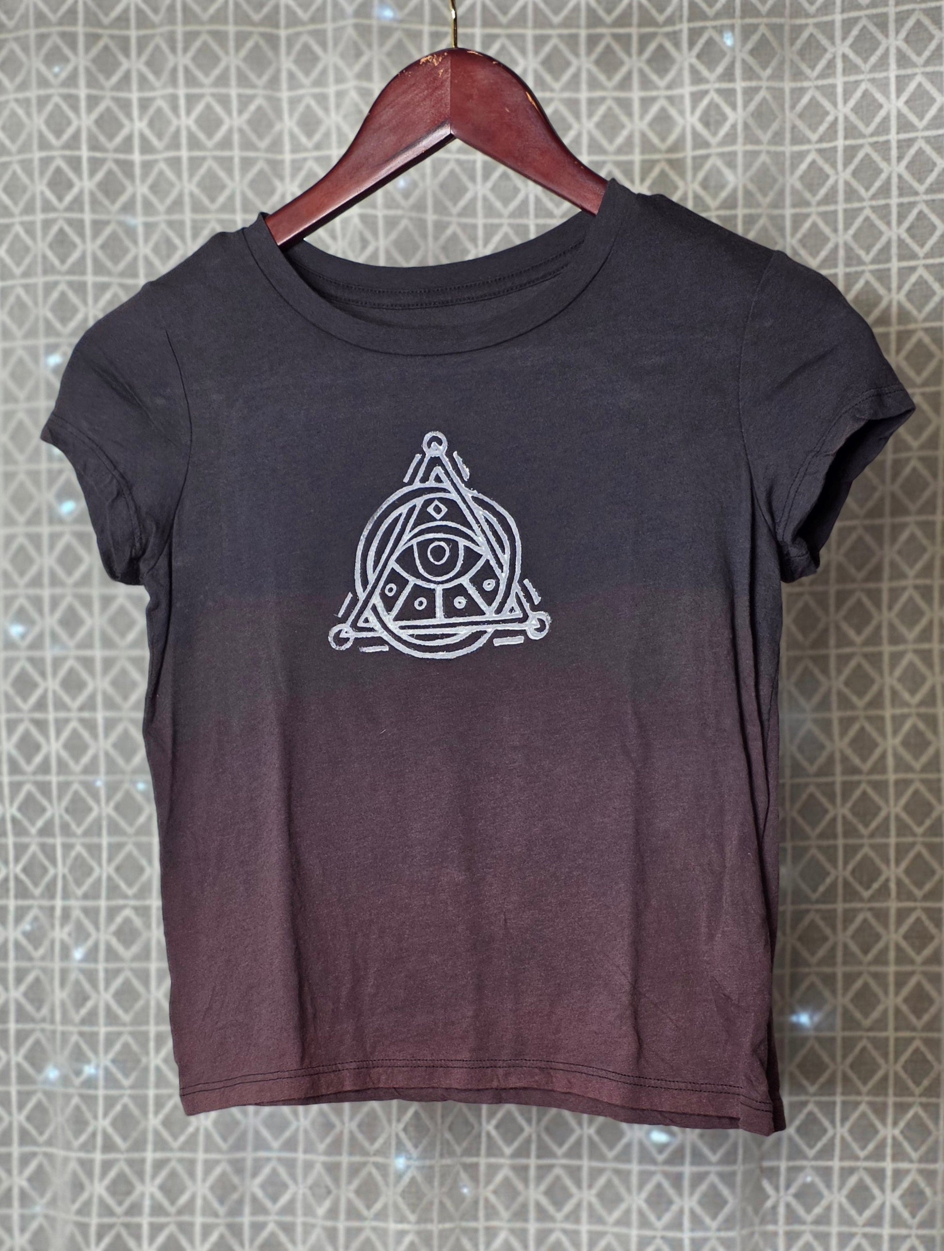 Black t-shirt with a white triangle symbol on a hanger against a patterned wall.