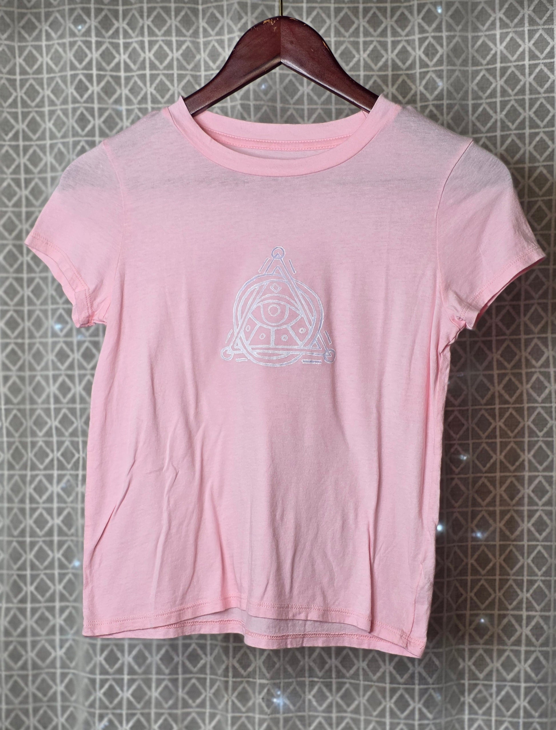 Pink t-shirt with a white logo on a hanger against a patterned wall.