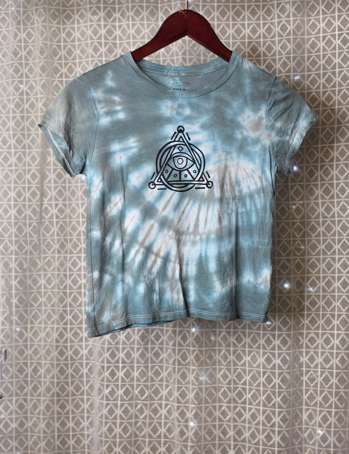 Tie-dye t-shirt with a symbol on a hanger against a patterned wall.
