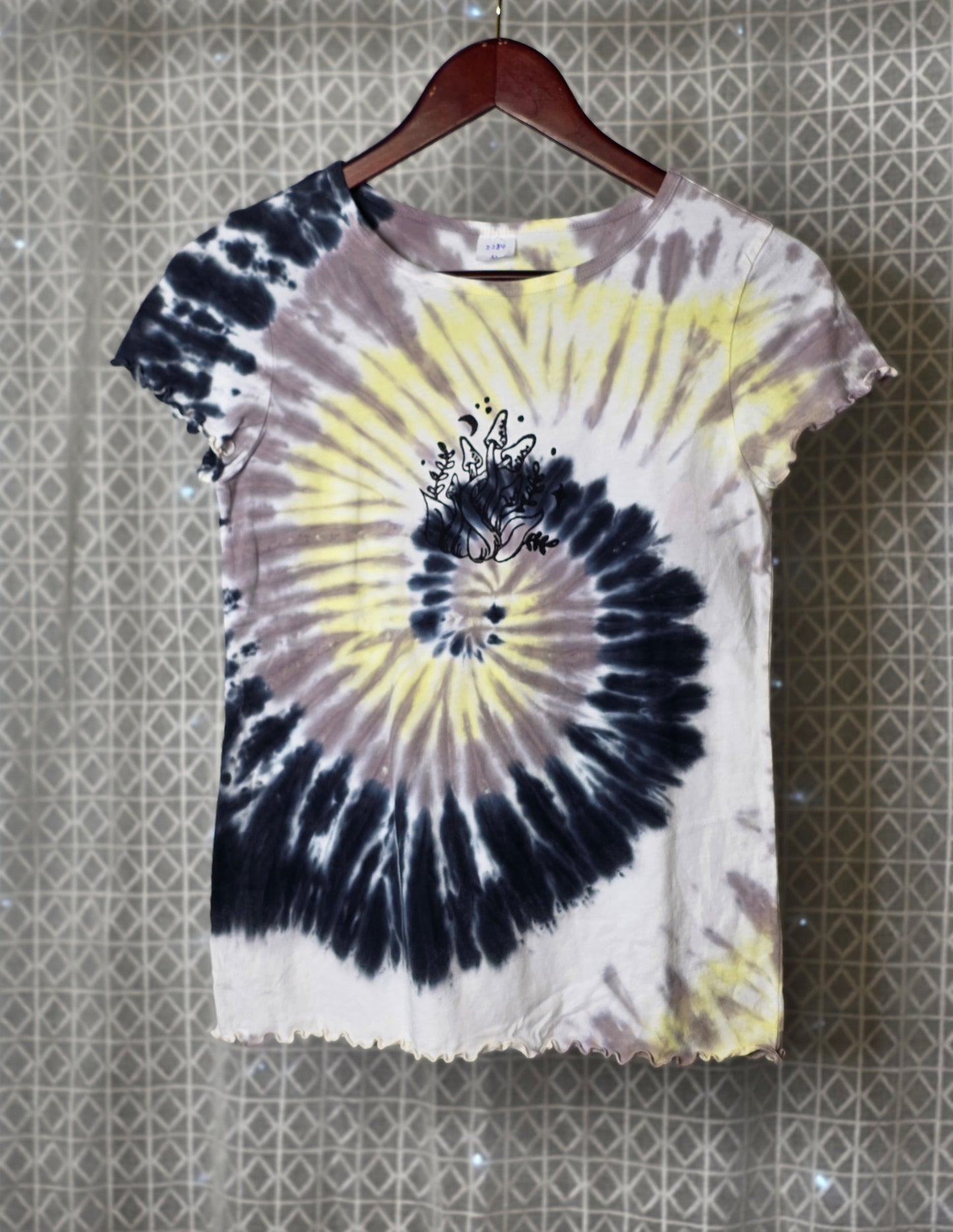 Tie-dye t-shirt with a spiral design on a hanger against a patterned wall.