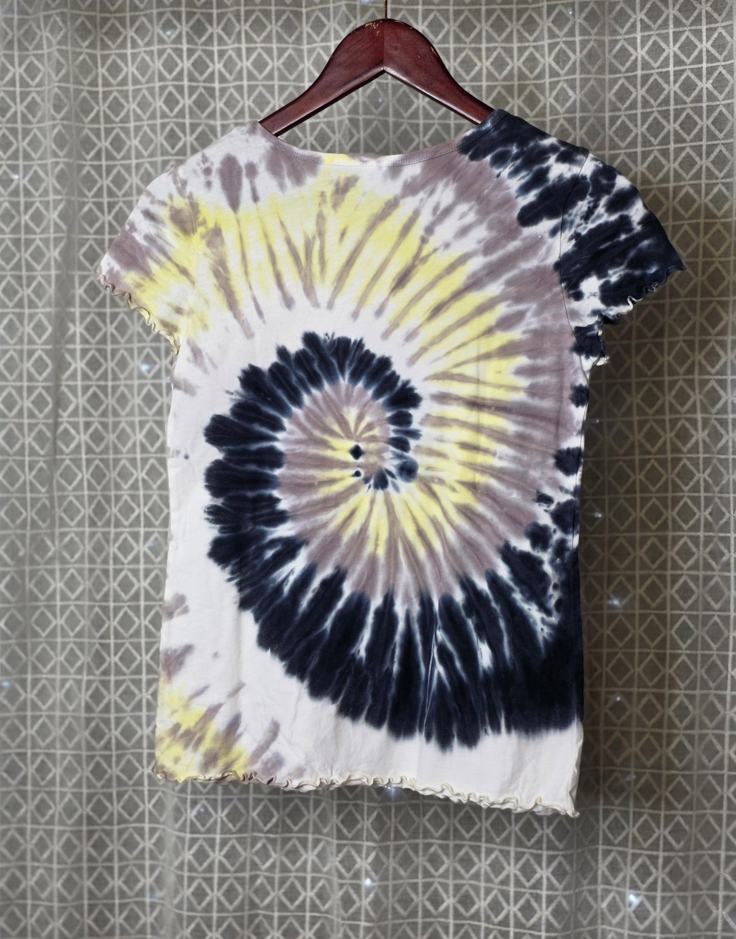 Tie-dye t-shirt with a spiral pattern on a hanger against a textured wall.