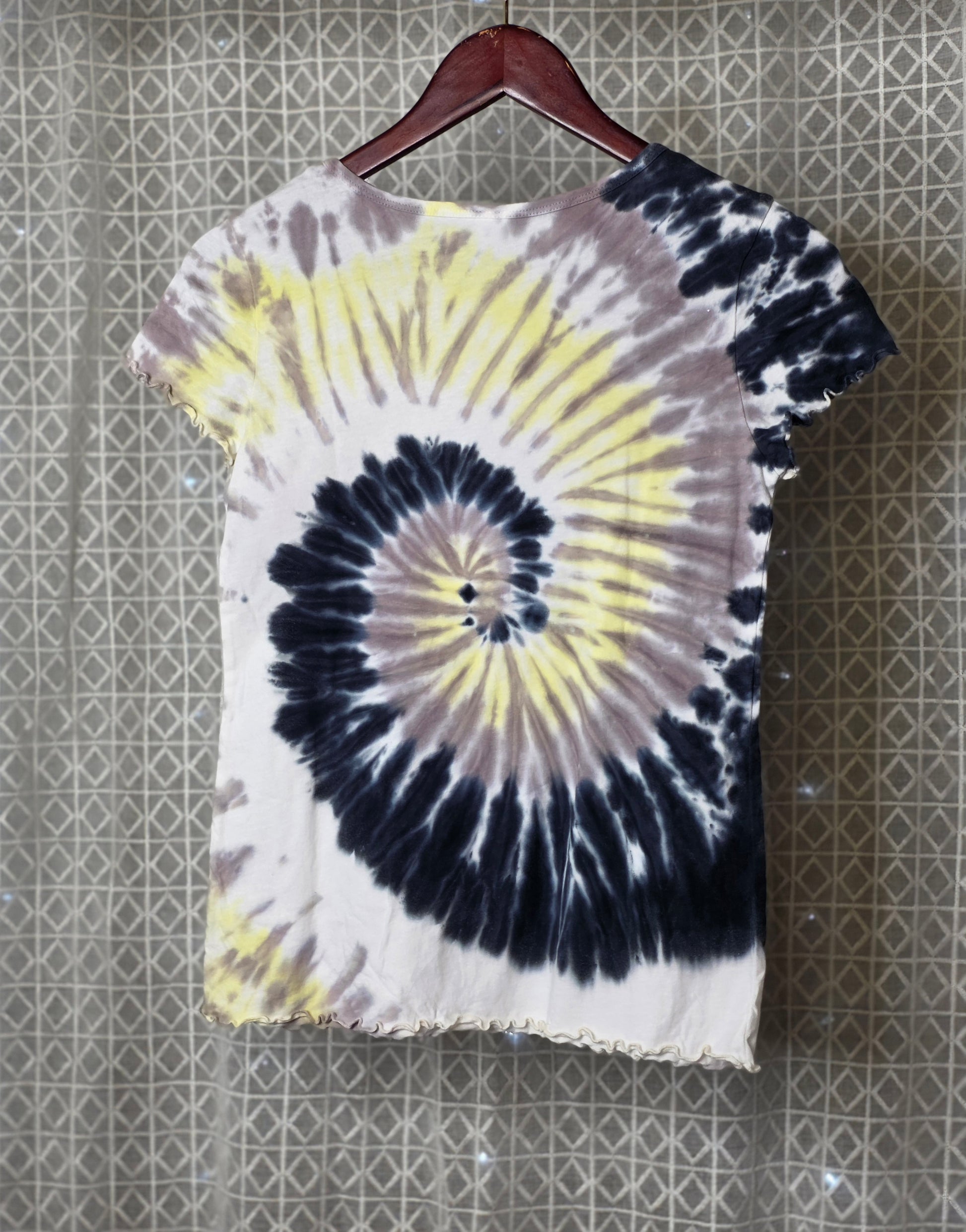Tie-dye t-shirt with a spiral pattern on a hanger against a textured wall.