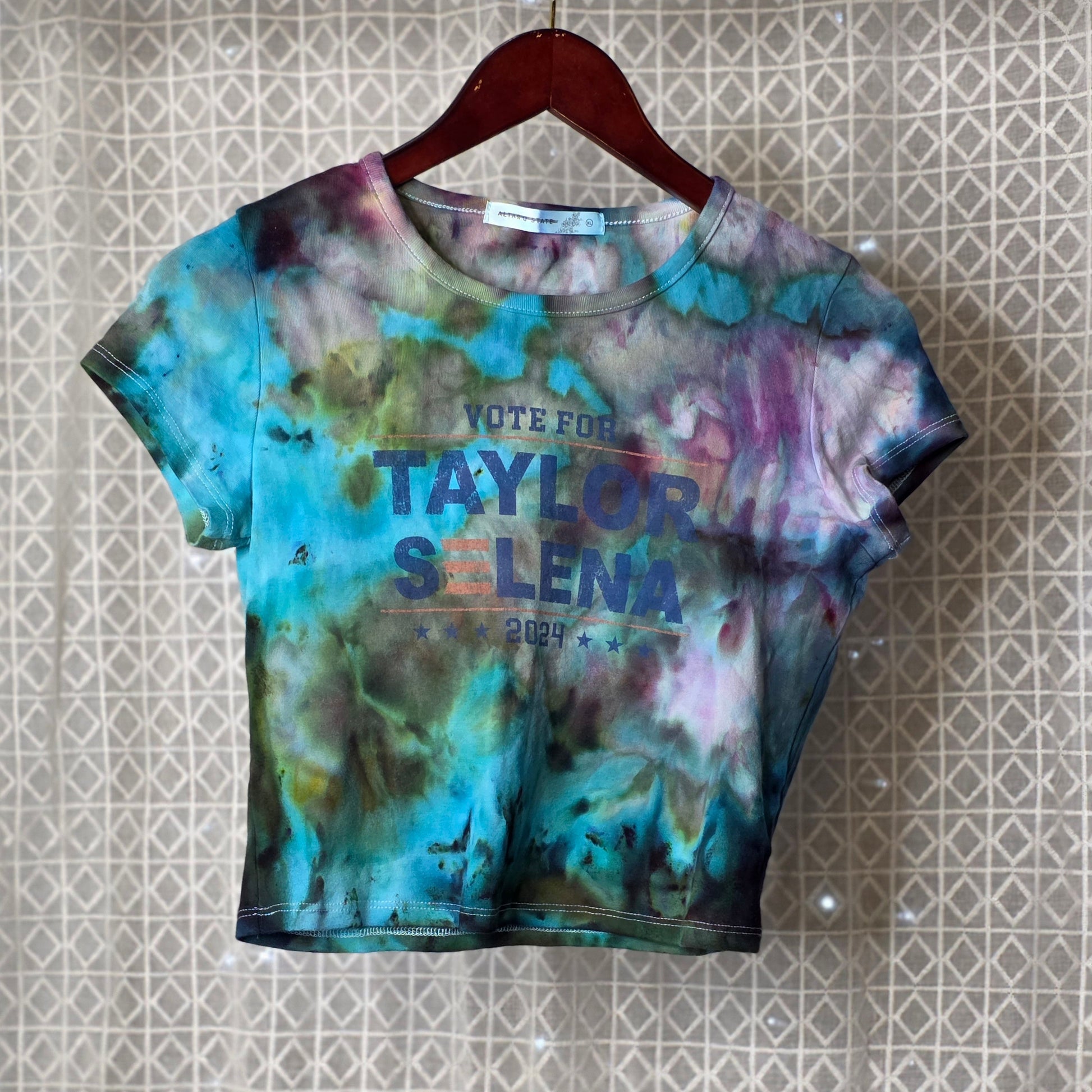 Tie-dye t-shirt with text on a hanger against a patterned wall