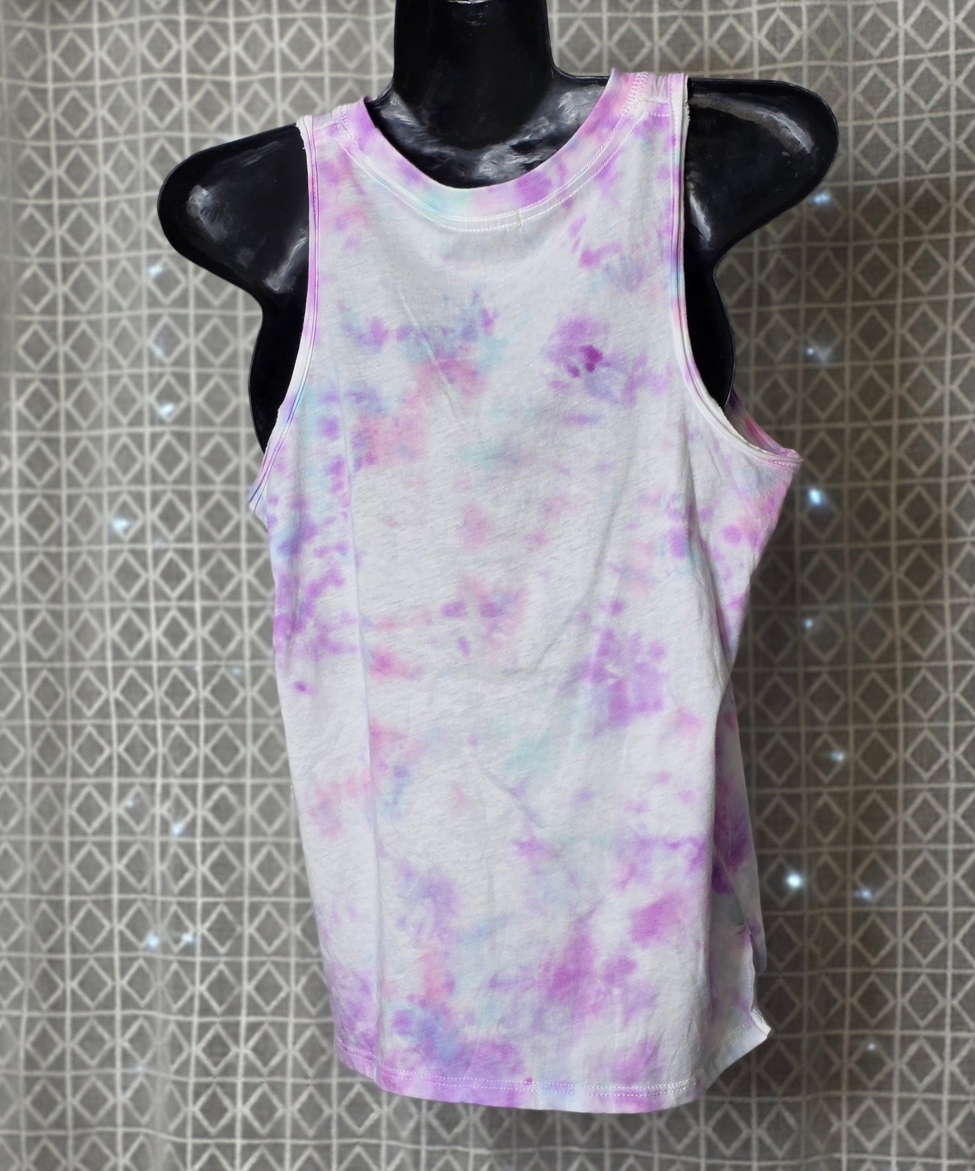 White tank top with purple and pink watercolor design on a mannequin against a patterned wall.