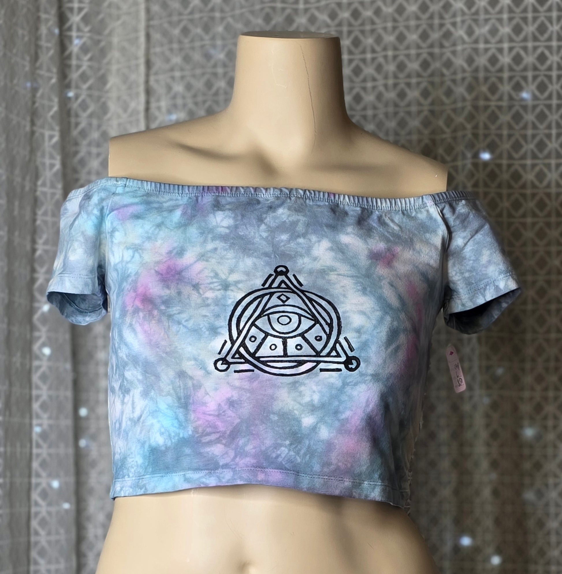 Tie-dye off-shoulder top with a symbol design on a mannequin against a patterned wall.