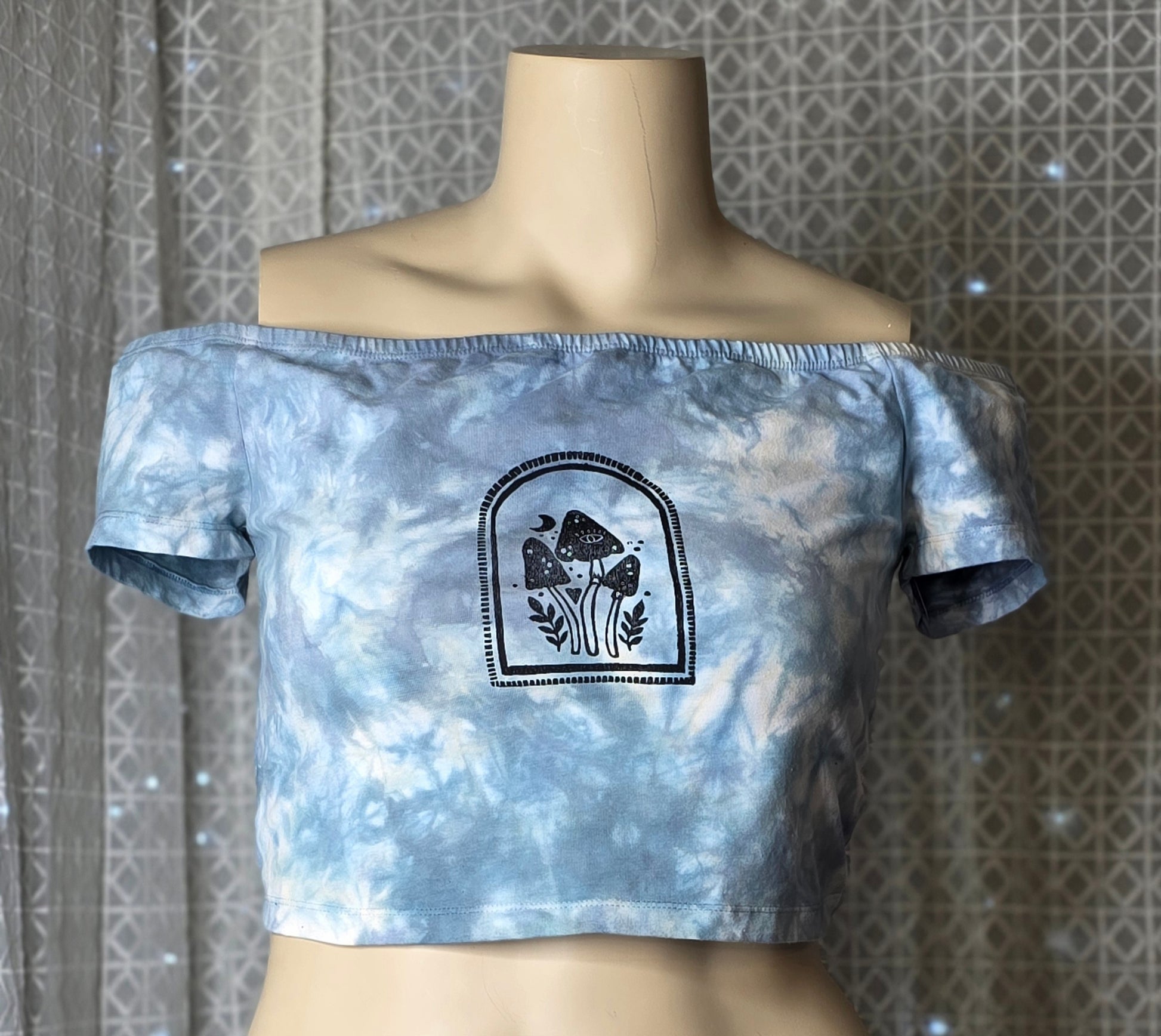 Blue tie-dye crop top with a black graphic design on a mannequin against a patterned wall.
